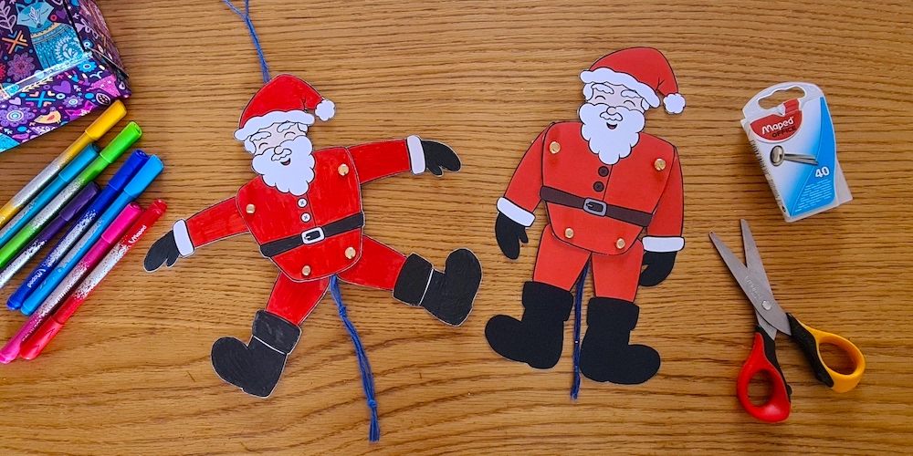 Two Santa puppets lay on a desk with felt tip pens and scissors