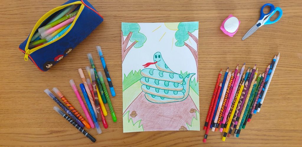 A piece of paper with a drawing of a snake laying on a desk surrounded by stationery