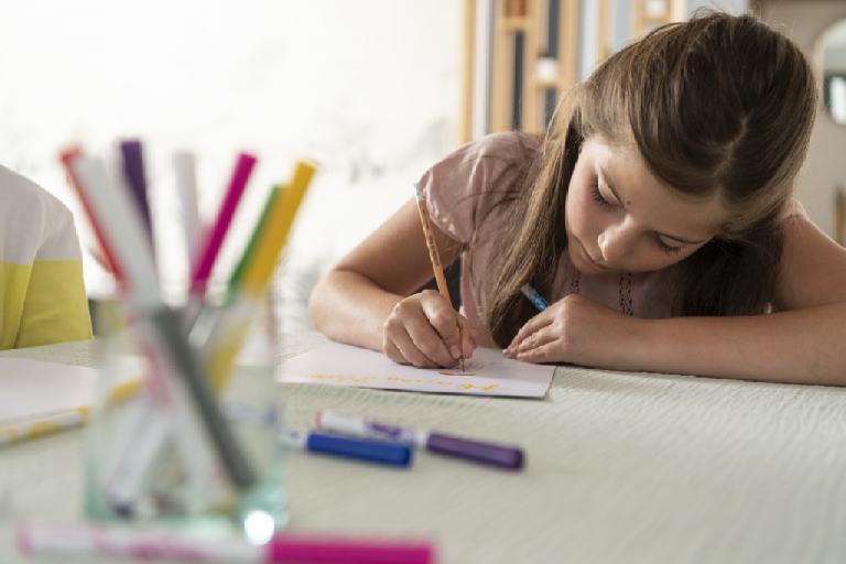 A girl stat at a table drawing a picture, surrounded by colouring pencils and felt pens